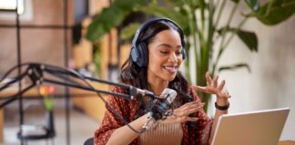 Podcast 2025 starten: Video Podcast & KI clever nutzen Junge Frau sitzt am Tisch und nimmt einen Podcast auf vor Ihr stehet ein Lapptop und ein Großes Podcast Mikrophon