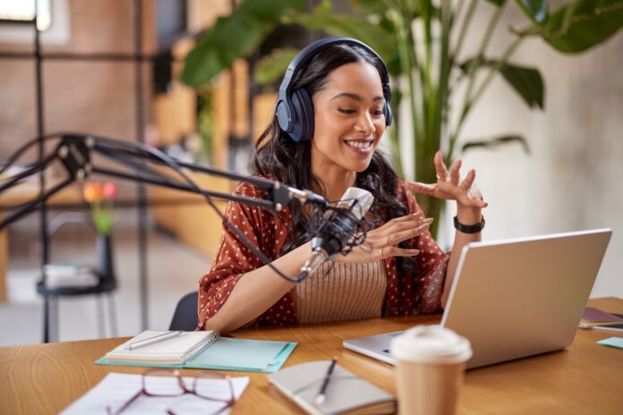 Podcast 2025 starten: Video Podcast & KI clever nutzen Junge Frau sitzt am Tisch und nimmt einen Podcast auf vor Ihr stehet ein Lapptop und ein Großes Podcast Mikrophon