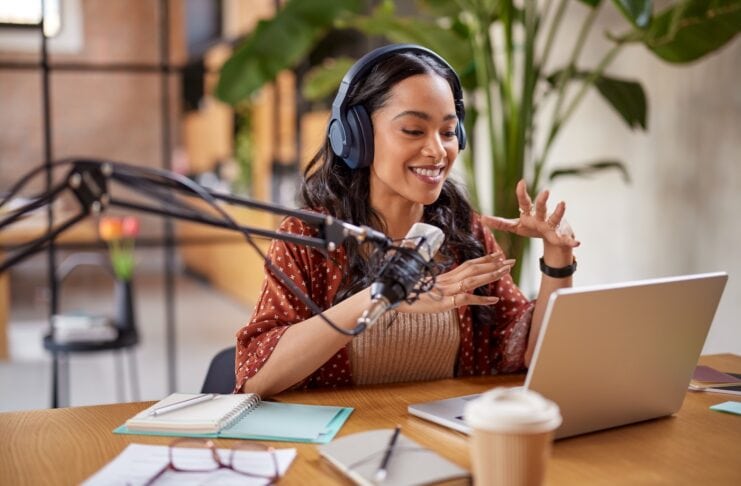 Podcast 2025 starten: Video Podcast & KI clever nutzen Junge Frau sitzt am Tisch und nimmt einen Podcast auf vor Ihr stehet ein Lapptop und ein Großes Podcast Mikrophon