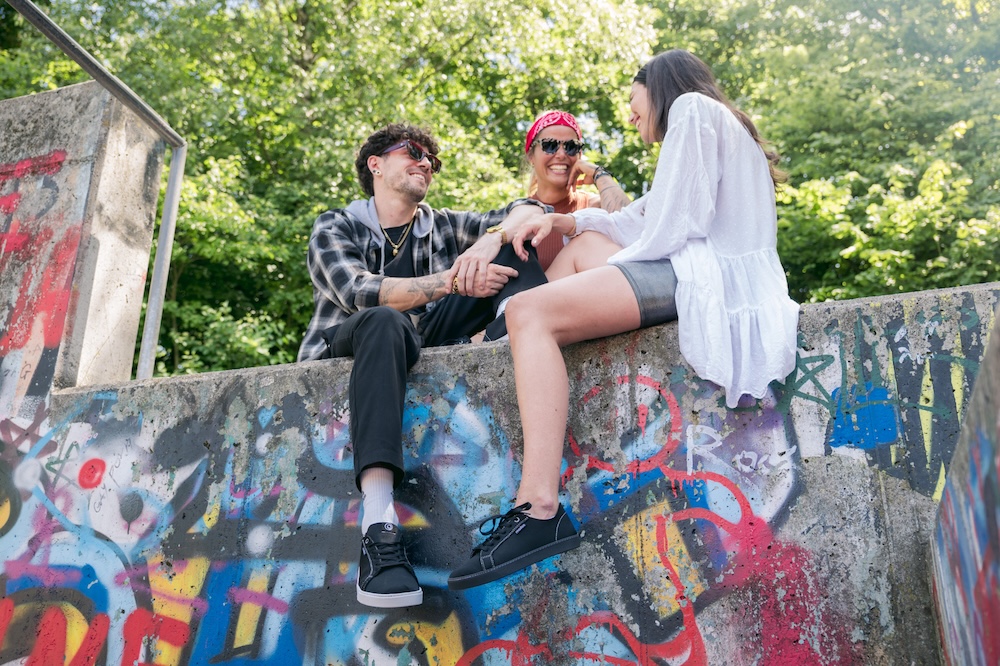 Madoo – Schuhe und Sneaker für echte Kreislaufwirtschaft 3 menschen auf einer mauer sitzend schuhe zeigend