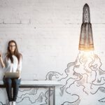 Startup im Nebenerwerb: Chancen, Risiken, rechtliche Aspekte und Praxistipps für eine erfolgreiche Gründung neben dem Hauptberuf. Junge Frau sitzt auf Tisch mit einem Lapptop auf den Beinen und im Hintergrund ist einen WQeisse Backsteinwadn auf die eine Rakete gemalt ist
