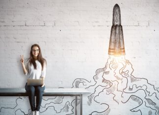 Startup im Nebenerwerb: Chancen, Risiken, rechtliche Aspekte und Praxistipps für eine erfolgreiche Gründung neben dem Hauptberuf. Junge Frau sitzt auf Tisch mit einem Lapptop auf den Beinen und im Hintergrund ist einen WQeisse Backsteinwadn auf die eine Rakete gemalt ist